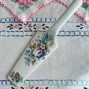 May include: A white ceramic cake server with a floral design featuring blue, yellow, and purple flowers. The server has a long handle and a gold trim. The server is on a white embroidered tablecloth.