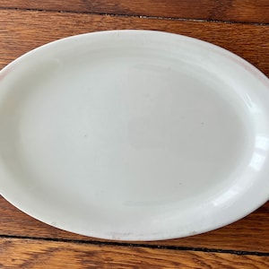 May include: A white oval serving platter. The ceramic dish has a smooth surface and a slightly raised edge. The platter is set against a wooden background, showcasing its clean design and simple elegance. Ideal for serving appetizers or main courses.