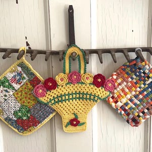 May include: Three handmade pot holders hanging on a vintage rake head. The pot holders are square and feature patchwork designs, crochet basket with floral accents, and woven patterns. Colors include yellow, red, blue, and green.