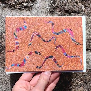 May include: A greetings card with a design of colourful snakes on an orange background. The snakes have striped patterns in shades of pink, blue, and yellow. The card is held against a textured stone wall.
