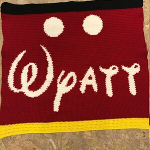 May include: Crocheted blanket with a red background, white Mickey Mouse ears, and the words "Disney" in white. The blanket has a yellow border.
