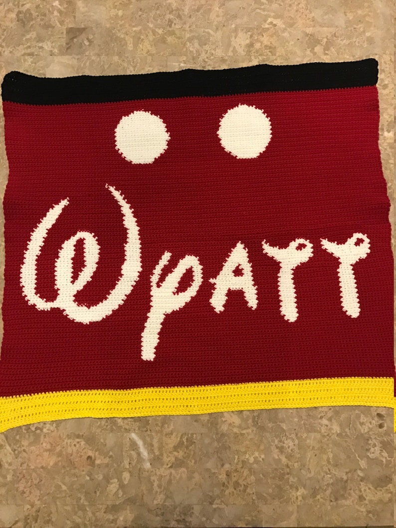 May include: Crocheted blanket with a red background, white Mickey Mouse ears, and the words "Disney" in white. The blanket has a yellow border.
