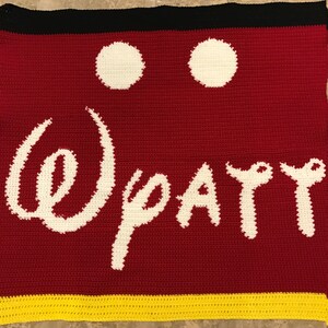 May include: Crocheted blanket with a red background, white Mickey Mouse ears, and the word "Disney" in white. The bottom border is yellow.