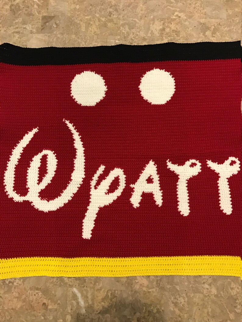 May include: Crocheted blanket with a red background, white Mickey Mouse ears, and the word "Disney" in white. The bottom border is yellow.