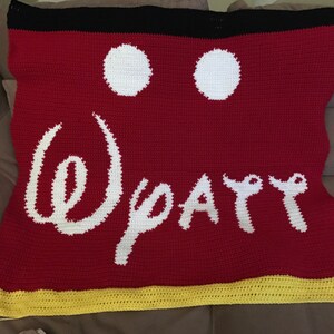 May include: Red crocheted blanket with a white Mickey Mouse head design and the words "Disney" in white. The blanket has a yellow border.