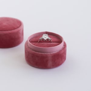 Puede incluir: Una caja de anillo con un forro de terciopelo en color rosa polvoriento. La caja está abierta y un anillo de plata con una gran piedra preciosa transparente está dentro.