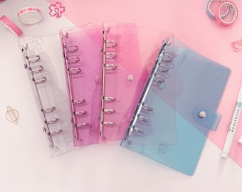 May include: Four clear plastic binders with different coloured translucent covers. The binders are open and show the rings for holding papers. The covers are light blue, pink, light purple, and clear.