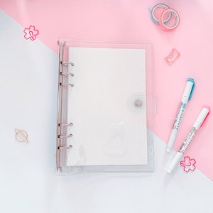 Può includere: Un raccoglitore in plastica trasparente con un inserto di carta bianca a pois. Il raccoglitore ha sei anelli e una chiusura a bottone in plastica trasparente. Il raccoglitore è su una superficie rosa.