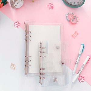 May include: Two clear plastic ring binders with silver rings. The larger binder has a white interior and the smaller binder has a white interior with the text "HANA" printed on it. The binders are on a pink surface with various decorative items.