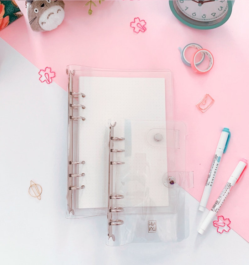 May include: Two clear plastic ring binders with silver rings. The larger binder has a white interior and the smaller binder has a white interior with the text "HANA" printed on it. The binders are on a pink surface with various decorative items.