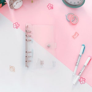 May include: A clear plastic binder with six silver rings and a silver button closure. The binder is on a pink and white surface with a few decorative items.