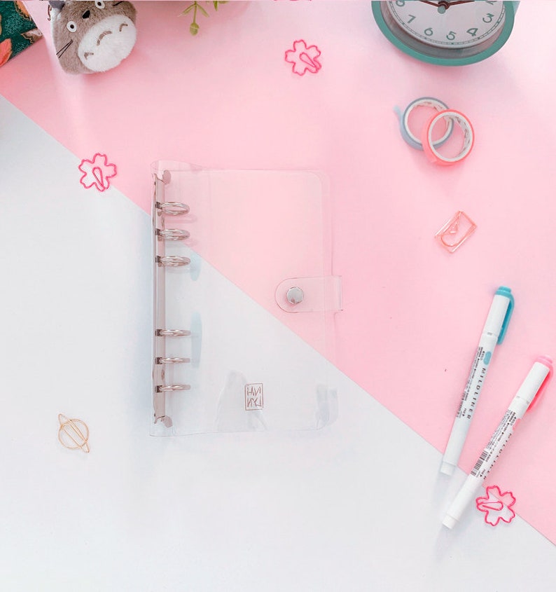 May include: A clear plastic binder with six silver rings and a silver button closure. The binder is on a pink and white surface with a few decorative items.