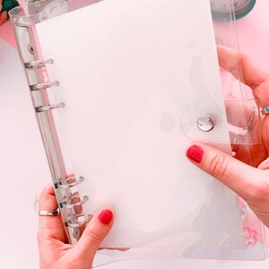 May include: A clear plastic binder with a white paper insert and silver rings. The binder has a silver button closure.