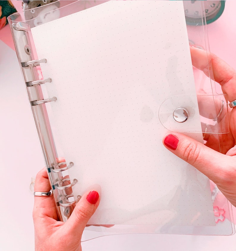 May include: A clear plastic binder with a white paper insert and silver rings. The binder has a silver button closure.