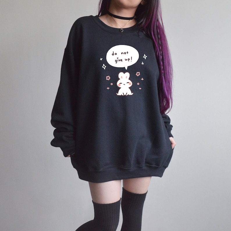 K&ouml;nnte beinhalten: Schwarzes Sweatshirt mit einer wei&szlig;en Sprechblase mit dem Text "do not give up!" und einer Cartoon-Hasen-Grafik. Der Hase ist wei&szlig; mit rosa Akzenten. L&auml;ssiger Stil.