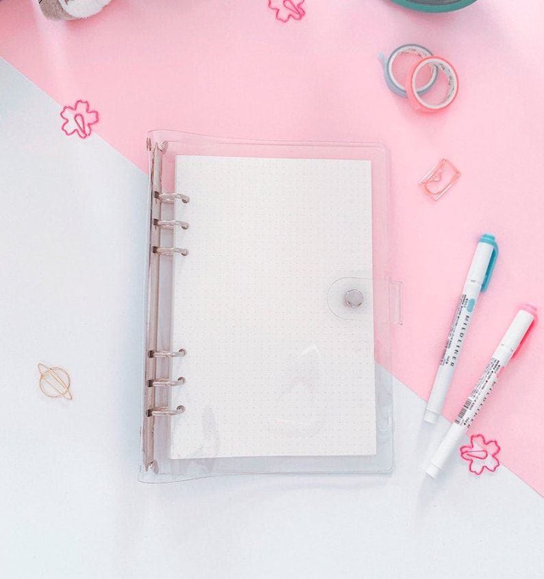 May include: A clear plastic binder with a white, dotted paper insert. The binder has six rings and a clear plastic button closure. The binder is on a pink surface.