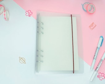 May include: A clear plastic binder with a white gridded paper insert. The binder has six silver rings and a red elastic closure. The binder is on a pink and white surface.