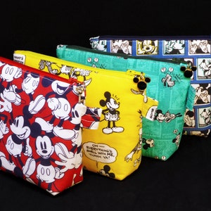 May include: Five different Mickey Mouse themed zippered pouches. The pouches are made of fabric and feature various Mickey Mouse cartoon images. The pouches are lined up in a row from largest to smallest. The largest pouch is blue with a white Mickey Mouse image. The next pouch is green with a white Mickey Mouse image. The next pouch is yellow with a white Mickey Mouse image. The next pouch is red with a white Mickey Mouse image. The smallest pouch is red with a white Mickey Mouse image.