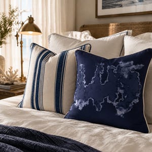 May include: Two square throw pillows on a bed. One pillow features a cream background with vertical navy blue stripes. The other pillow is solid navy blue with a white, cloud-like abstract pattern and white piping along the edges.