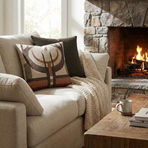 Può includere: Un divano beige con cuscini decorativi e una coperta lavorata a maglia. Un cuscino presenta un motivo geometrico marrone, rosso e beige. Un cuscino marrone scuro è dietro. Un camino acceso e un tavolino di legno sono sullo sfondo.