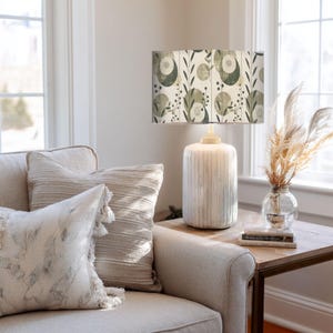 May include: A cream-colored ceramic lamp with a patterned lampshade featuring green and white botanical designs. The lamp sits on a wooden side table next to a sofa with decorative pillows. A vase of dried pampas grass adds a natural touch.