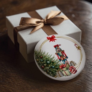May include: A white, round Christmas ornament with a watercolor illustration of a nutcracker, Christmas tree, and wrapped gifts. The ornament is framed by a gold line and a red bow. A white gift box with a brown ribbon is in the background.