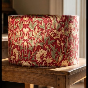 May include: A cylindrical drum lampshade featuring a deep red background with a repeating botanical pattern of light green leaves and cream-colored floral blossoms. The fabric has a visible woven texture throughout the entire surface.