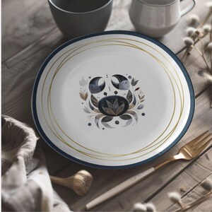 Op de afbeelding: Witte keramische plaat met een blauwe rand en gouden accenten. De plaat heeft een krabontwerp met maandetails en bloemelementen. Een gouden vork en een houten tafel zijn op de achtergrond.
