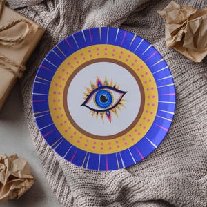 May include: A decorative plate with a blue, yellow, and white design. The plate features a central eye motif with a blue iris and intricate details. The border is blue with white lines, and the plate has a diameter of approximately 10 inches.