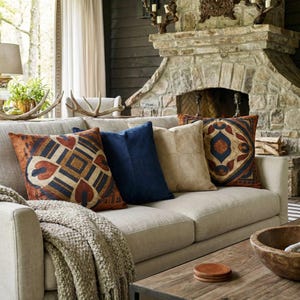 May include: A beige sofa with several decorative pillows in shades of blue, tan, and rust. The pillows feature geometric and patterned designs. A knitted throw blanket and a wooden coffee table complete the cozy living room setting.