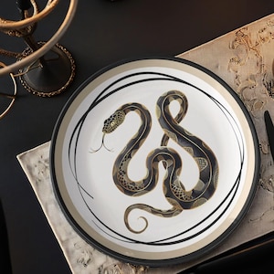 Puede incluir: Un plato decorativo con un diseño de serpiente en negro, dorado y blanco. La serpiente está enrollada y detallada con escamas y una lengua bífida. El plato tiene un borde negro con detalles dorados y blancos.