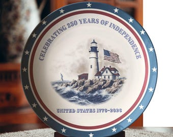 May include: A decorative plate commemorating 250 years of United States independence. The plate features a lighthouse, American flag, and the Statue of Liberty. The rim is blue with white stars and red stripes. Text reads "Celebrating 250 Years of Independence" and "United States 1776-2026".