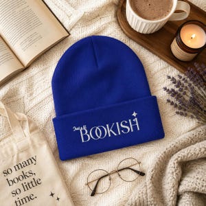 May include: A royal blue beanie with the embroidered text "Just a lil BOOKISH" in white. The beanie is folded at the brim. A tote bag with the text "so many books, so little time" is also visible.
