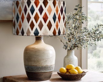 May include: A table lamp with a cylindrical drum shade featuring a repeating diamond pattern in shades of orange, brown, and dark blue. The lamp base is a rounded, cream-colored ceramic vessel with a textured, dark brown bottom section.