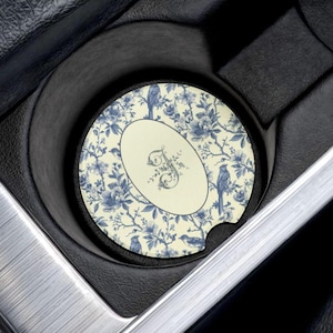 Pode incluir: Uma base de copo redonda para carro com um design floral e de pássaros em azul e creme. A base de copo apresenta um centro oval com uma inicial ornamentada. A base de copo está colocada num porta-copos de carro.
