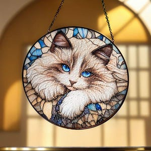 A stained glass window hanging with a round frame and a ragdoll cat with blue eyes. The cat is looking to the right and has a brown nose. The background is a mosaic of blue, brown, and white glass pieces.
