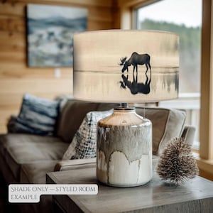 May include: A decorative lamp with a cylindrical lampshade featuring a silhouette of a moose drinking water. The lamp base is a ceramic design with a white and brown glaze. The text "SHADE ONLY - STYLED ROOM EXAMPLE" is at the bottom.
