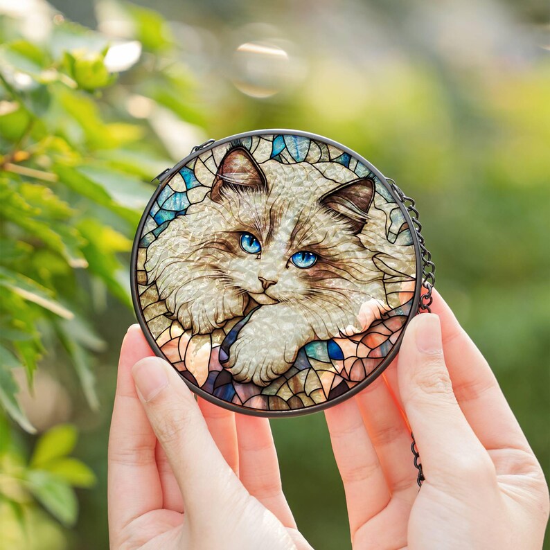 a small round stained glass image of a bicolor ragdoll cat face , large and fluffy coat with big paws and vibrant blue eyes. it is being held in a woman's hands outside in the sunlight