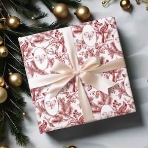 Puede incluir: Un regalo envuelto con un patrón de toile de Jouy rojo y blanco con pájaros y follaje. El regalo está atado con una cinta y un lazo de satén rosa claro. Adornos navideños dorados y vegetación en el fondo.