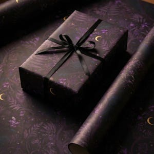 May include: A rectangular gift box wrapped in dark purple paper featuring a botanical pattern of ferns and leaves with gold crescent moon accents, tied with a black satin ribbon. Beside it are two rolls of the same matching dark purple wrapping paper.