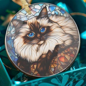 Puede incluir: Adorno navideño redondo con un gato de ojos azul brillante y pelaje marrón y blanco. El gato está sobre un fondo de estilo vidriera con tonos azules, blancos y naranjas. Se adjunta una cinta dorada para colgar.