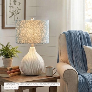 May include: A white ceramic table lamp with a patterned lampshade. The lampshade has a light blue background with a white floral design. The lamp sits on a wooden table next to a small potted plant and a stack of books. A blue blanket is draped over a chair.