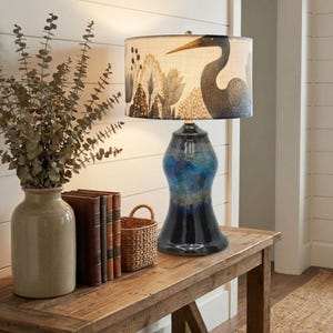 May include: A decorative table lamp with a blue and black ceramic base and a patterned lampshade. The lampshade features a heron design with a forest background. The lamp is placed on a wooden table with a vase of greenery, books, and a small basket.