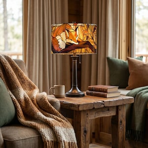 May include: A decorative table lamp with a stained glass shade featuring a fox design in shades of orange, brown, and cream. The lamp has a dark metal base and sits on a rustic wooden side table. A cozy scene with a blanket and books.