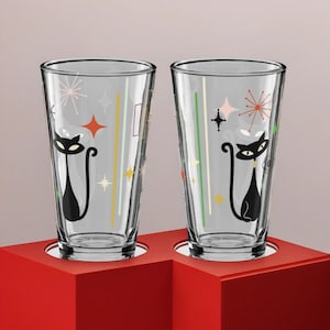 Könnte beinhalten: Zwei klare Glasbecher mit schwarzen Katzensilhouetten und geometrischen Mustern im Retro-Stil. Die Gläser stehen auf einer roten Oberfläche.