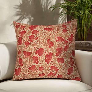 Puede incluir: Cojín cuadrado con un estampado floral. El cojín presenta un diseño repetitivo de flores y hojas rojas sobre un fondo crema. El cojín está colocado sobre un sofá blanco, con una planta en el fondo.