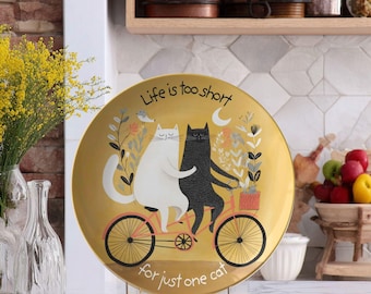 May include: A round decorative plate with a gold background featuring a white cat and a black cat riding a red tandem bicycle together. The text "Life is too short for just one cat" is printed in black script around the cats and the bicycle.