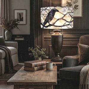 May include: A decorative table lamp with a stained glass shade featuring a black crow perched on a branch. The lamp has a dark bronze base and is placed on a wooden table. The room has a cozy, vintage aesthetic.