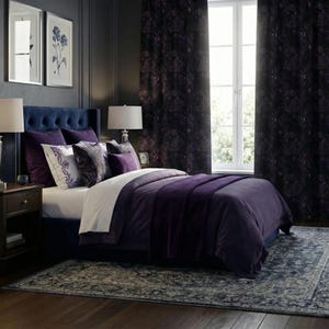 A bedroom scene featuring a bed with a navy blue headboard and a plush, deep purple comforter. Decorative pillows in shades of purple and white adorn the bed. Matching purple curtains frame a window, and a patterned rug lies on the floor.
