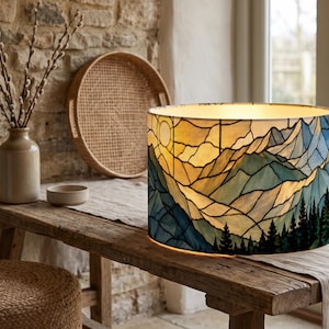 May include: A cylindrical lamp shade featuring a stained glass style design of a mountain landscape at sunset. The pattern includes layered blue and gold peaks, a bright yellow sun, and a dark green forest of pine trees along the bottom edge.